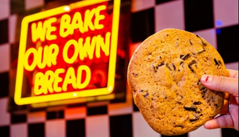 A chocolate chip cookie held up in front of a sign that reads, "We Bake Our Own Bread"